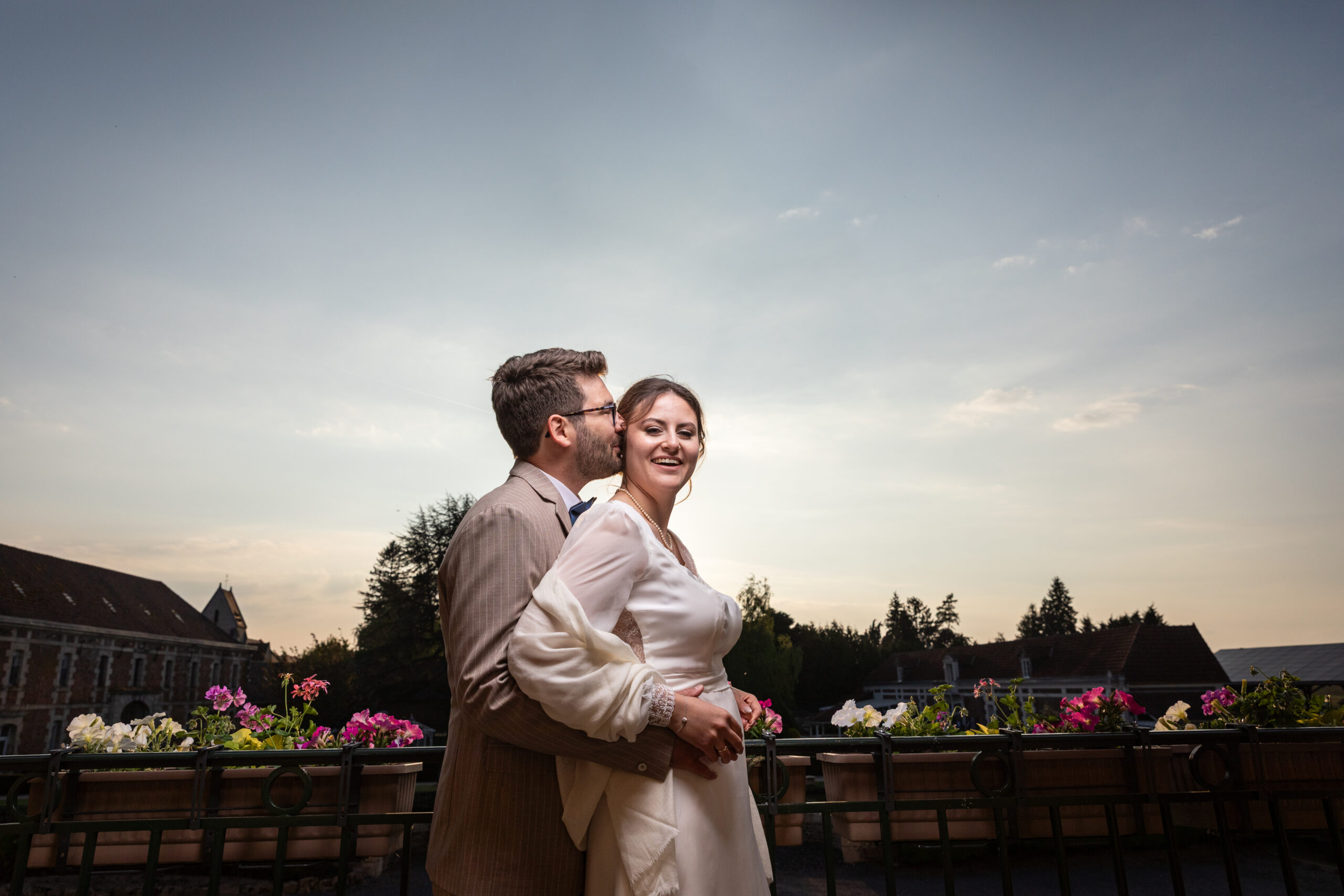 un homme embrassent sa femme sur la Joux sur un balcon.l'homme porte un costume marron et la femme en robe beige claire.