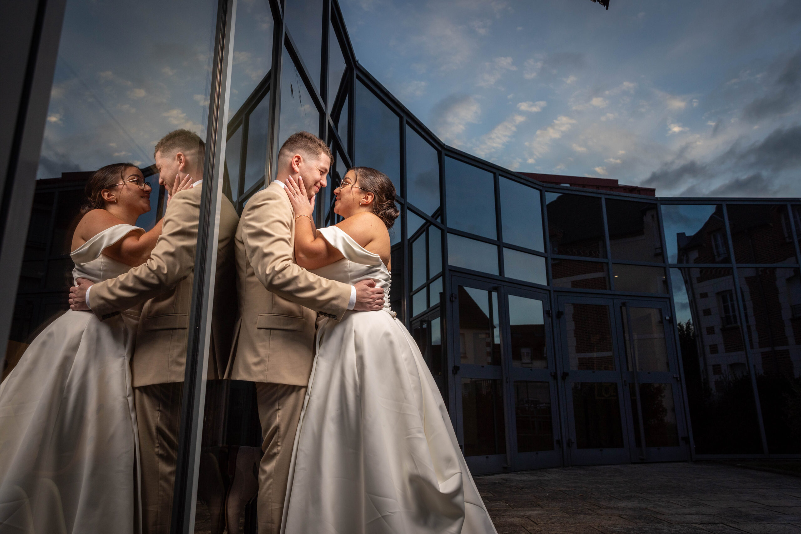 un couple de marier s'enlacent tendrement devant une façade en verre qui reflets leur reflet ainsi que celui du soleil qui ce couche.
